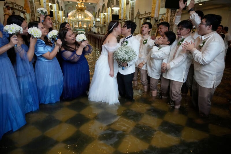 A bride and groom kiss inside a flooded church, surrounded by smiling bridesmaids in blue dresses and groomsmen in barong tagalog, all standing in ankle-deep water and celebrating joyfully.