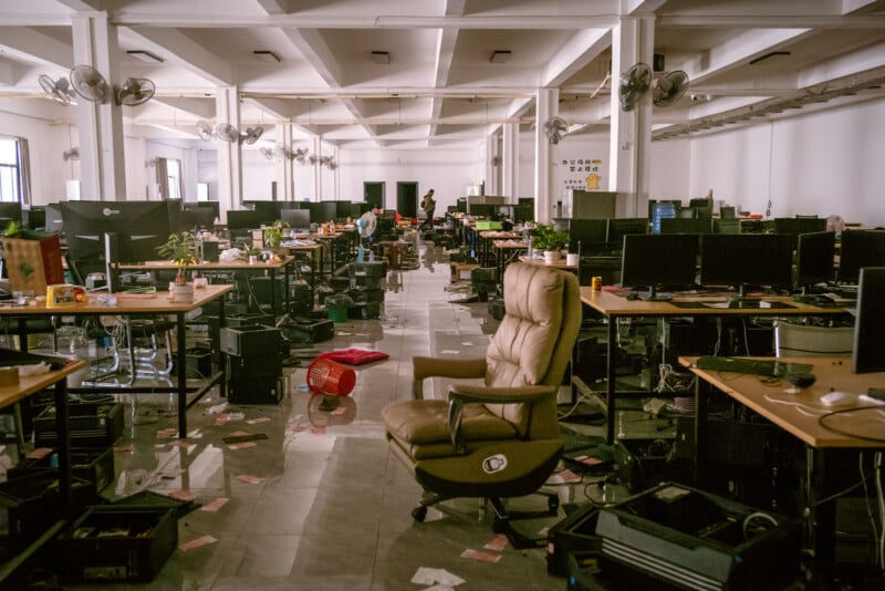 A spacious office with many empty desks, scattered papers, and chairs in disarray. Computer monitors are on the desks, and the area appears abandoned and messy, with some items on the floor.