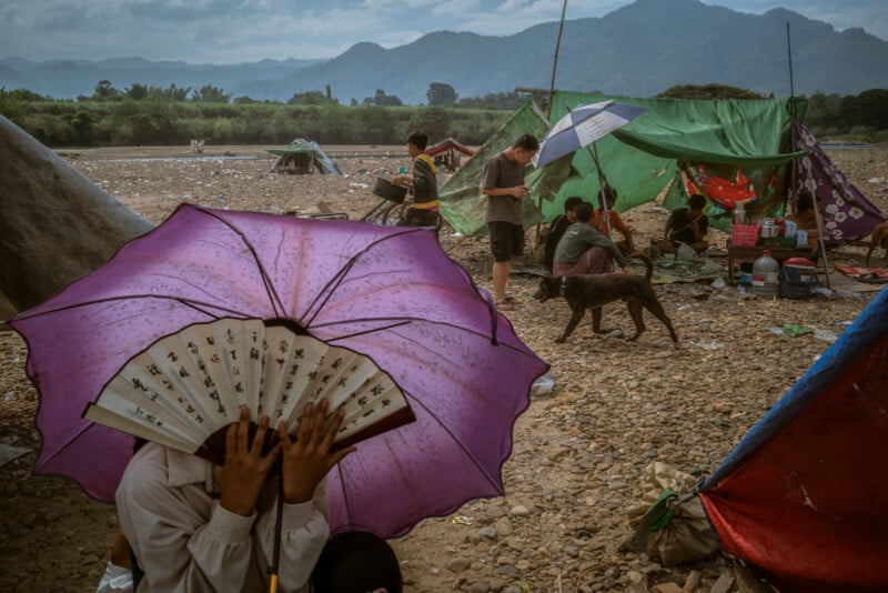People gather at makeshift tents on a rocky riverbank with mountains in the background. In the foreground, a person holds a purple umbrella and fan, partially covering their face. A dog and scattered belongings are visible.