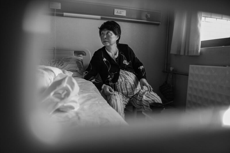 A woman in pajamas sits on a hospital bed, looking to the side. The scene is framed by a blurred foreground, and the room appears dimly lit with light coming from a window.