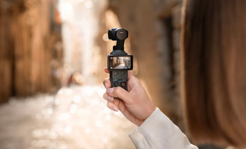 A person holds a handheld camera with a small display screen, capturing a narrow, sunlit street with blurred, warm-toned buildings in the background.