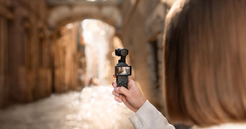 A person with shoulder-length hair holds a small handheld camera, capturing video in a sunlit, narrow alley with stone walls and an arched bridge in the background. The camera screen displays the alley scene.