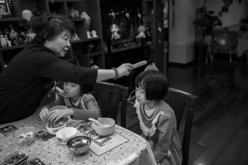 An older woman reaches over a dining table to touch the head of a young girl, while another child sits nearby and prepares food. The scene is set inside a cozy, decorated home.