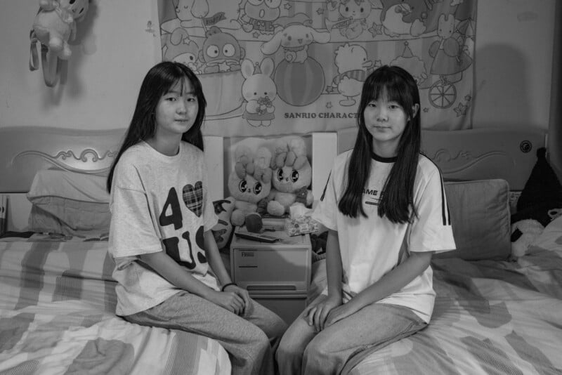 Two young girls with long hair sit side by side on a bed in a bedroom decorated with Sanrio character items, plush toys, and a cartoon-themed tapestry. The scene is in black and white.