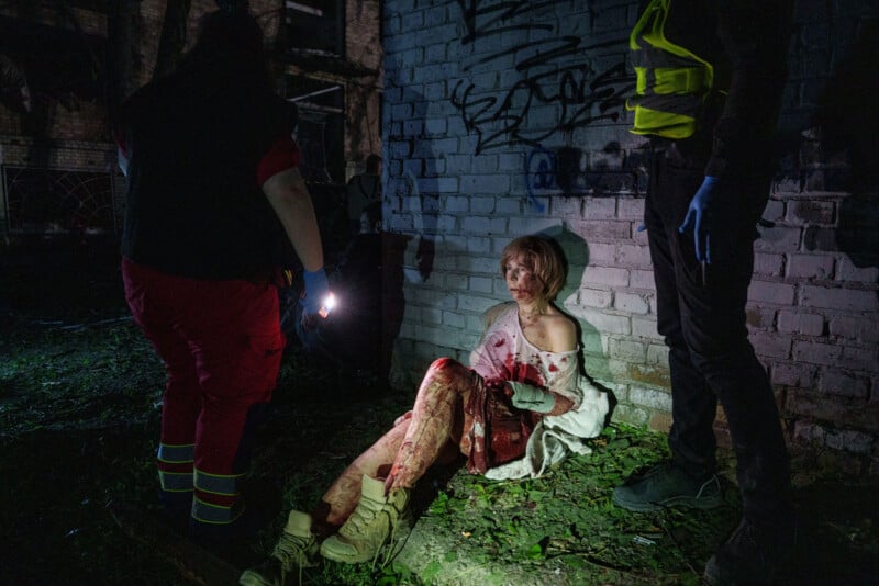 A bloodied woman in a white shirt sits on the ground against a graffiti-covered brick wall at night, surrounded by two emergency responders, one shining a flashlight on her.