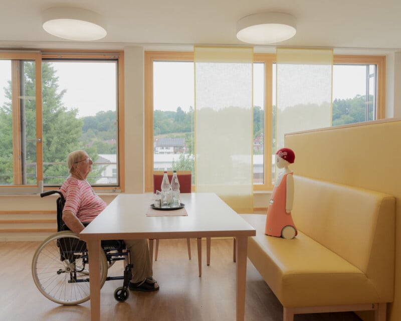 An elderly person in a wheelchair sits at a table across from a small humanoid robot on a yellow bench in a bright room with large windows and light wood flooring.