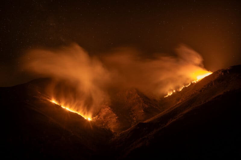 A nighttime view of a wildfire spreading across mountain slopes, with bright orange flames and thick smoke rising into the dark sky, partially illuminated by the fire.