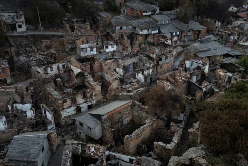 Aerial view of a village with many houses severely damaged or destroyed, showing collapsed roofs, burnt walls, and debris, indicating the aftermath of a fire or disaster. Some buildings remain partially intact.