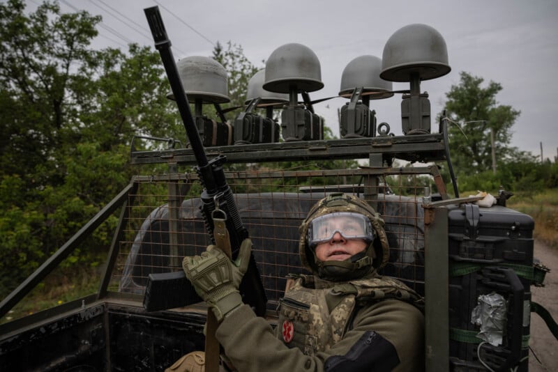 A soldier in military gear and goggles holds a rifle while sitting in a vehicle equipped with multiple electronic devices and antennas, surrounded by greenery on a cloudy day.