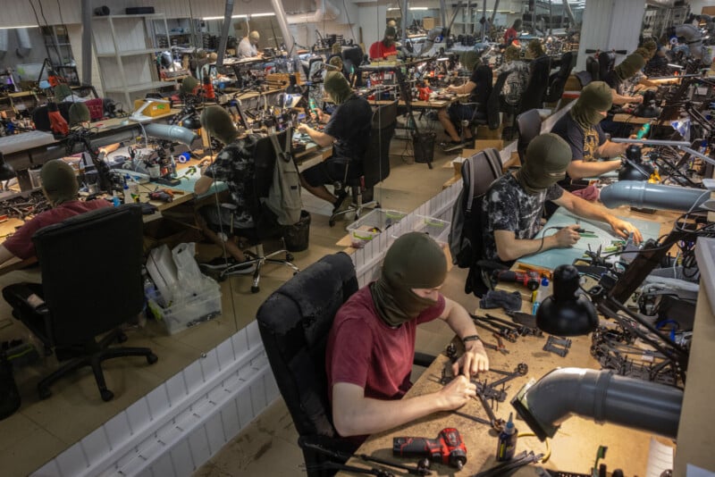 People wearing balaclavas work at individual desks in a crowded workshop, assembling or repairing small electronic devices or drones. The room is well-lit, with tools and equipment scattered on the tables.
