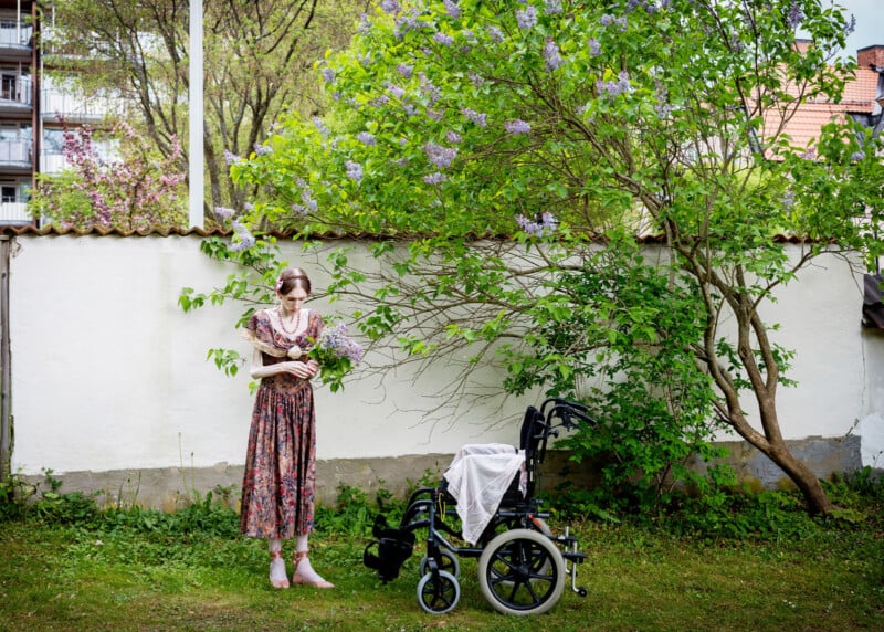 An elderly woman in a floral dress stands outdoors next to a wheelchair, holding lilac flowers beside a blooming lilac bush. A white cloth covers the wheelchair seat; buildings and a white wall are in the background.
