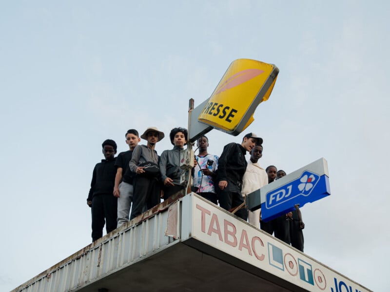 A group of people stand closely together on the roof of a building beneath a damaged "PRESSE" sign, with "TABAC LOTO" and "FDJ" signs visible on the building's edge, under a mostly clear sky.