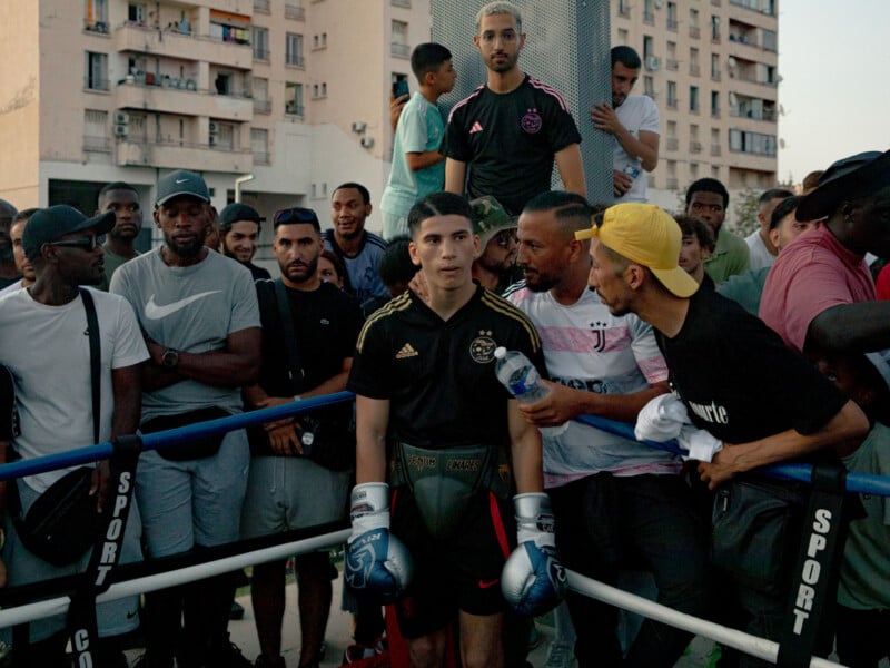 A young male boxer in gloves stands in a ring surrounded by a crowd of men outdoors; some are supporting and talking to him while others watch intently, with apartment buildings in the background.