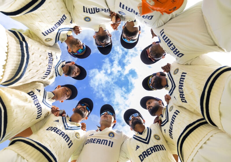 A group of cricket players in cream uniforms and caps stand in a huddle, looking up towards the camera, with blue sky and clouds visible above them. The word “DREAM11” is visible on their sweaters.