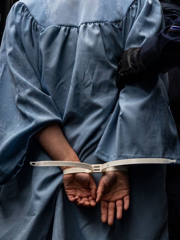 A person in a blue graduation gown stands with hands secured behind their back with white zip-tie handcuffs, while a gloved officer holds their arm.
