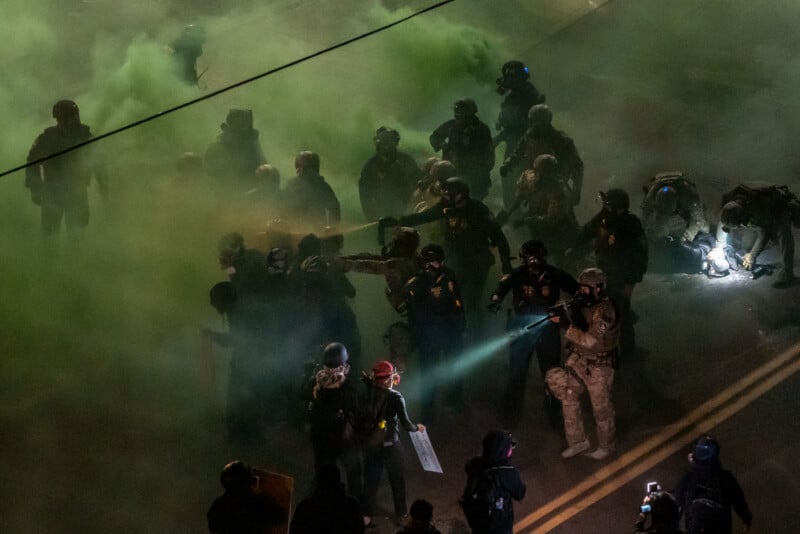 A group of law enforcement officers in riot gear stands amid green smoke on a street at night, facing a crowd of protesters. One officer shines a flashlight, while others kneel or stand in formation.