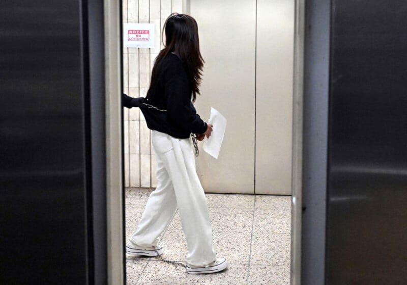 A person in a black sweatshirt and white pants holds papers while waiting in front of closed elevator doors in a building hallway. A notice is posted on the wall above the elevator.
