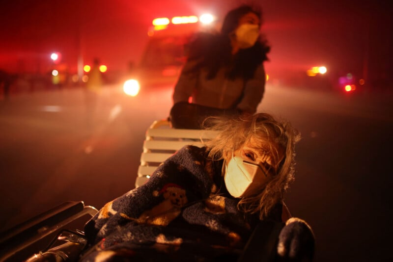 An elderly woman in a mask sits in a wheelchair, wrapped in a blanket, while another person pushes her. Emergency vehicle lights and smoke fill the background, creating an urgent and tense atmosphere.