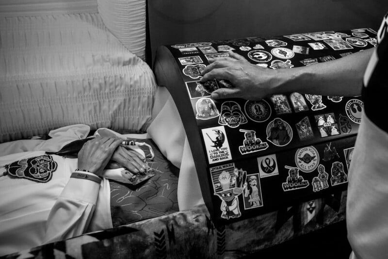 A person lies in a coffin with their hands clasped, wearing a suit and a wristband. Another person's hand rests on the coffin lid, which is covered with various colorful stickers. The image is in black and white.