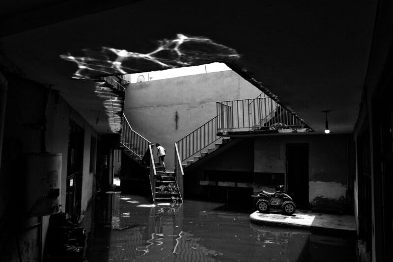 A black and white photo shows a flooded courtyard with a staircase leading up, a child walking up the stairs, and light reflections on the ceiling. A toy car sits on the water-covered ground.