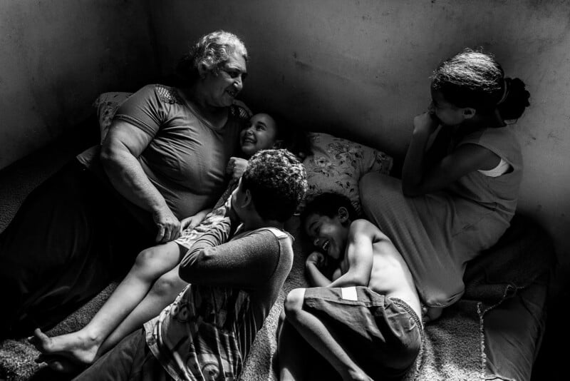 An older woman lies on a bed, smiling and laughing with four children around her. The group shares a joyful, intimate moment together in a simple, softly lit room.