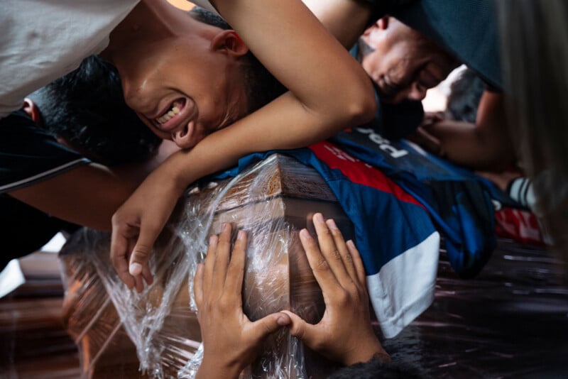 Several grieving people embrace and cry over a plastic-wrapped coffin, their faces contorted in sorrow as hands clutch the casket tightly in mourning.