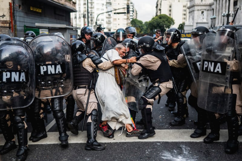 A man in white robes is pushed by a group of police officers in riot gear and shields marked "PNA" during a tense protest on a city street lined with buildings.