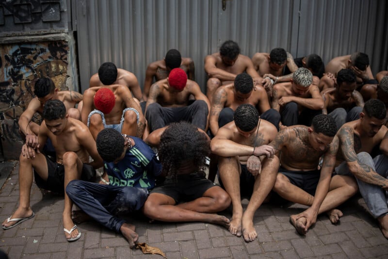 A group of shirtless men with bowed heads sit closely together on the ground against a metal fence, some with colored hair, appearing subdued or detained.