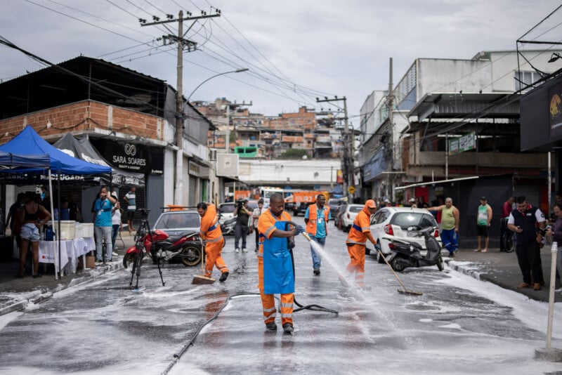 City workers in orange uniforms and blue aprons spray and scrub a street with soap and water. Bystanders and vendors watch from the sides, and buildings line the background of the urban scene.