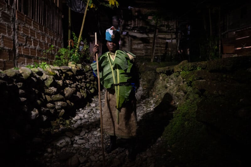 A person stands on a rocky path at night, wearing a traditional outfit made of leaves, a cloth skirt, and a blue and white headpiece, holding a long wooden stick, with stone walls and wooden houses in the background.