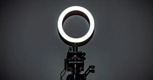 A brightly lit ring light is mounted on a black tripod stand against a dark, plain background.