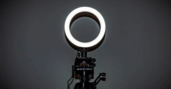 A brightly lit ring light is mounted on a black tripod stand against a dark, plain background.