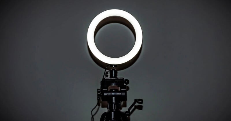 A brightly lit ring light is mounted on a black tripod stand against a dark, plain background.