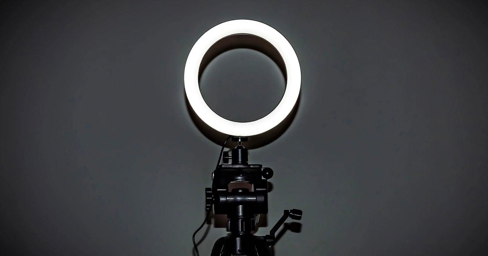 A brightly lit ring light is mounted on a black tripod stand against a dark, plain background.