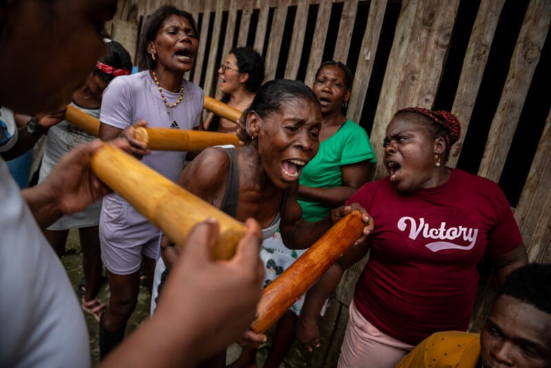 A group of women, some singing passionately and holding wooden sticks, gather closely together, expressing strong emotions near a wooden fence. One woman in a "Victory" shirt stands out in the foreground.