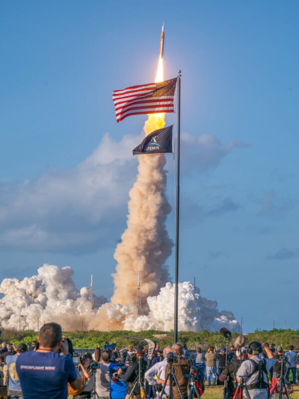 A rocket launches into a clear blue sky, leaving a trail of smoke as crowds of photographers and onlookers watch. An American flag and an Artemis mission flag are visibly flying in the foreground.