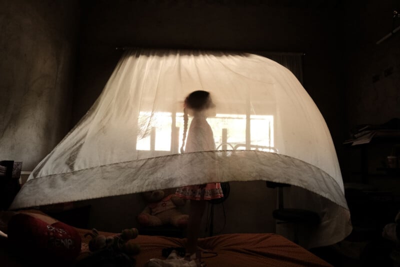 A young child stands on a bed behind a translucent mosquito net, softly backlit by sunlight streaming through a window in a dim room, creating a dreamy silhouette effect.