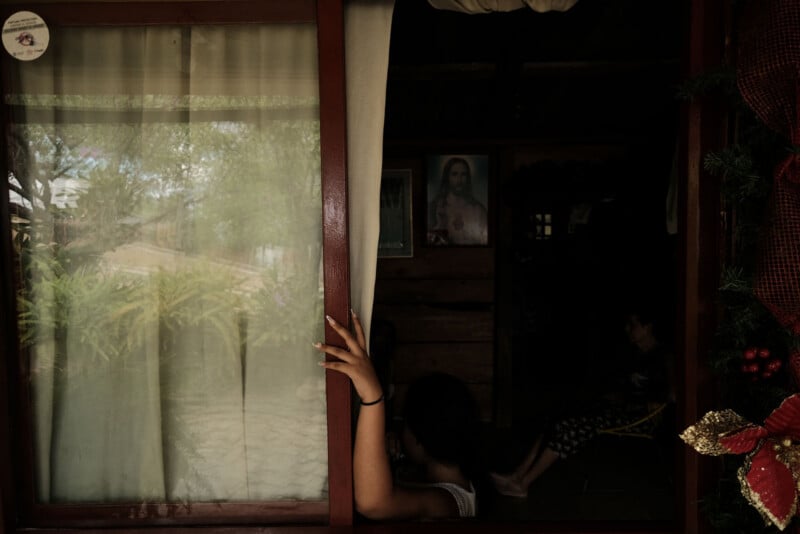 A person’s hand pulls back a curtain at a doorway, revealing a dimly lit room with another person sitting inside. Framed pictures and greenery are visible. The outdoor scene is partially reflected in the window.