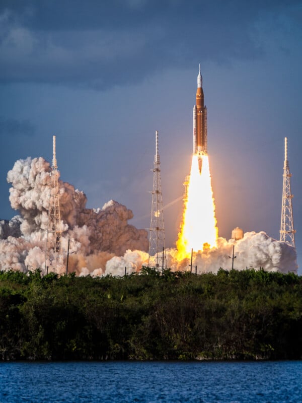 A rocket launches into the sky, emitting bright flames and thick clouds of smoke, surrounded by launch towers, with water and greenery in the foreground under a cloudy sky.
