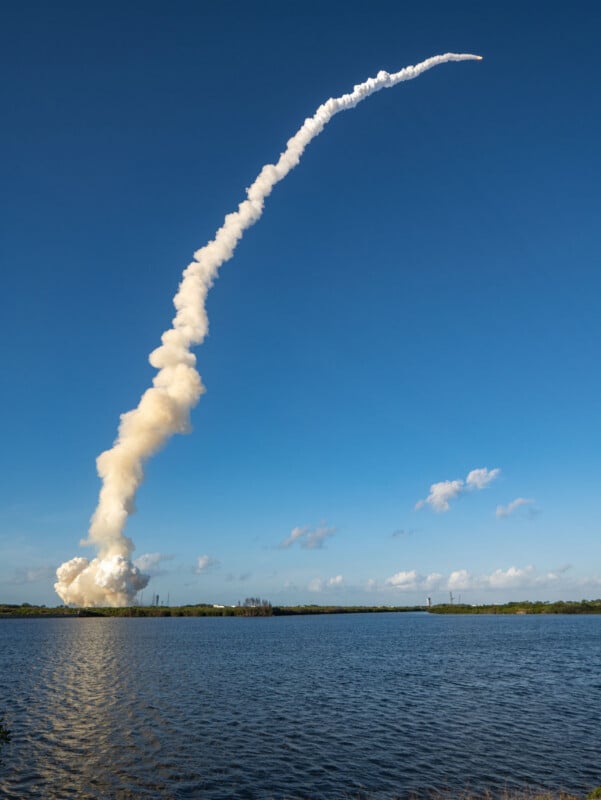 A rocket launches into a clear blue sky, leaving a thick trail of white smoke that curves upward over a body of water, with clouds and greenery visible on the horizon.