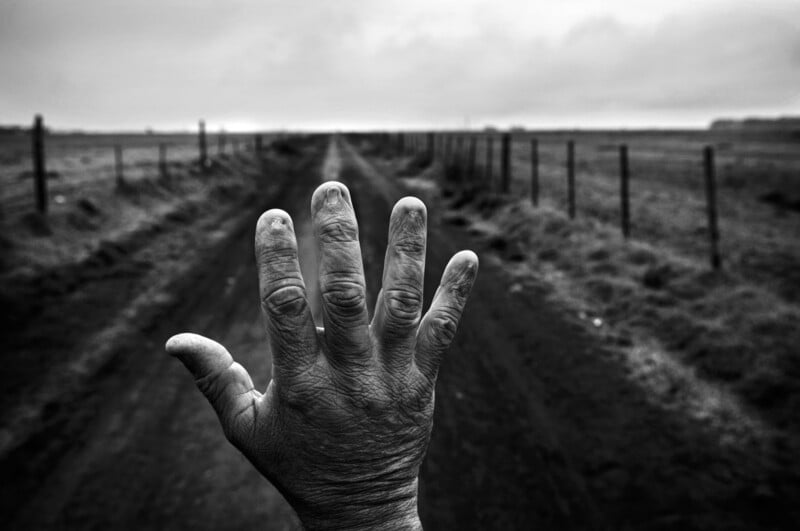 A close-up of a rough, weathered hand with fingers spread wide, held up against a rural dirt road flanked by fences, stretching into a cloudy, distant horizon. The image is in black and white.