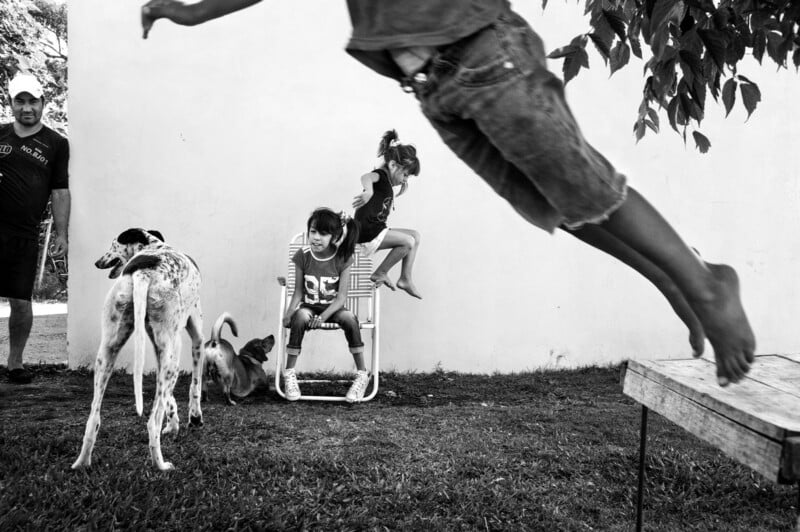 A playful outdoor scene shows a child mid-jump in the foreground, a dog and two children in the background, one sitting on a chair and another swinging. An adult stands to the side, all against a white wall on grass.