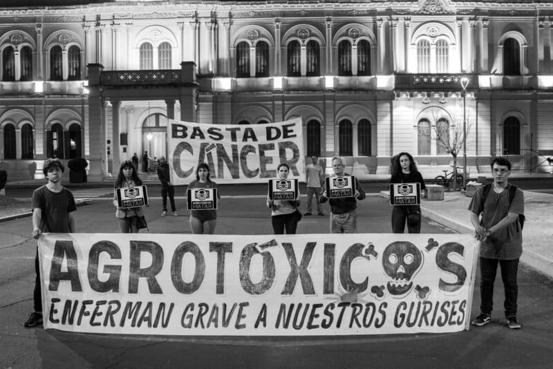 A group of people hold a large banner reading "AGROTÓXICOS ENFERMAN GRAVE A NUESTROS GURISES" with a skull, and a sign above reading "BASTA DE CÁNCER," in front of an ornate building at night.