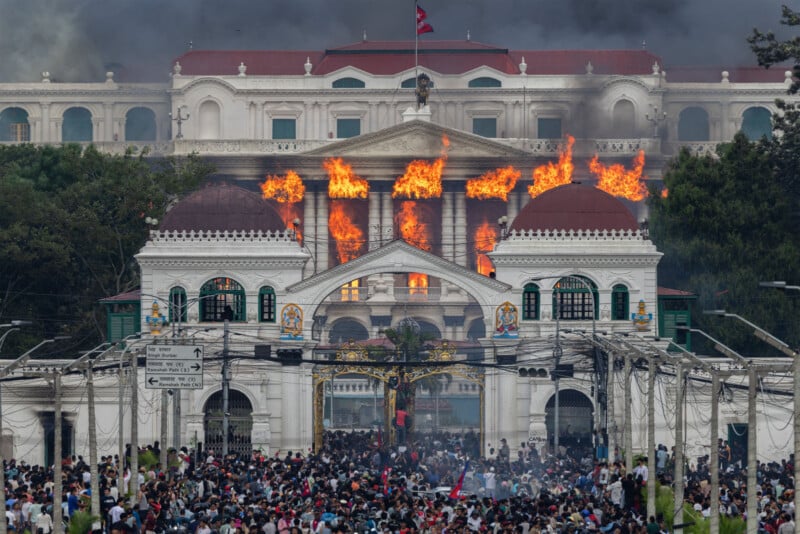A large crowd gathers in front of a grand white building with domes as flames and smoke pour from its upper windows, indicating a major fire.