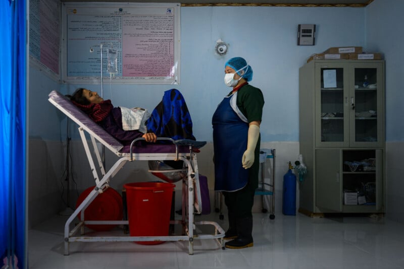 A patient lies on a hospital examination bed covered by a blanket while a healthcare worker in protective gear stands nearby in a medical clinic room with medical equipment and posters on the wall.