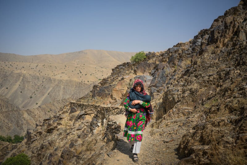 A woman in a colorful dress and headscarf carries a baby as she walks along a rocky mountain path. Arid hills and sparse greenery are visible in the background under a clear sky.