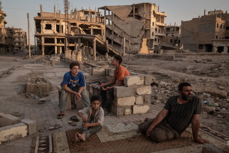 Four people, including three boys and a man, sit on a rug and rubble-strewn ground in front of heavily damaged, partially destroyed buildings in a war-torn urban area at sunset.