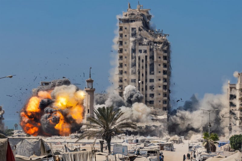 A large explosion engulfs the base of a high-rise building, sending flames, smoke, and debris into the air. The area below is filled with tents, palm trees, and power lines under a clear blue sky.