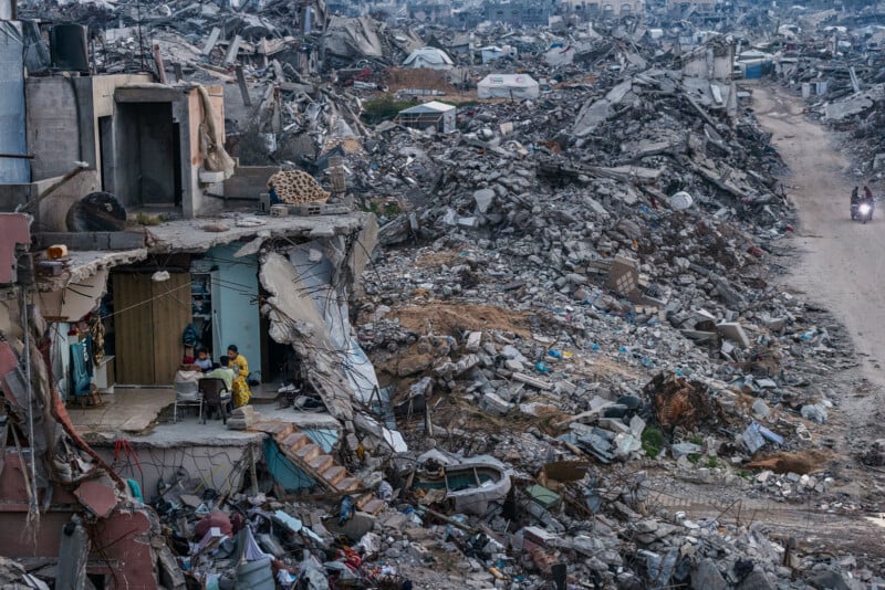 A family sits around a table in a partially destroyed building, surrounded by rubble and debris, while a dirt road stretches through the devastation with a motorcycle in the distance.