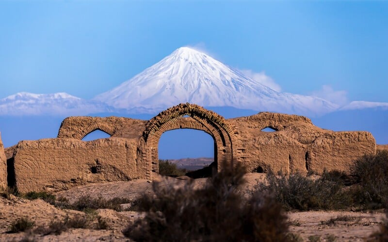 A snow-covered mountain rises in the background behind ancient, weathered brick ruins with an arched gateway, set in an arid landscape under a clear blue sky.